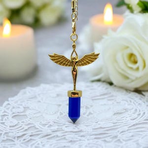 Peut inclure: Un pendentif en forme d'ange doré avec des ailes déployées, attaché à une pointe de cristal bleu. Le pendentif est suspendu à une chaîne et repose sur une surface en dentelle blanche.