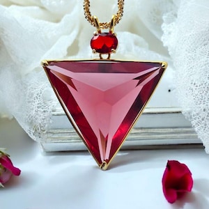 Peut inclure: Un collier doré avec un grand pendentif triangulaire rouge rubis. Le pendentif a un cadre doré et est suspendu à une fine chaîne dorée. Une petite gemme rouge de forme ovale est située au-dessus du triangle. Des pétales de rose sont éparpillés.