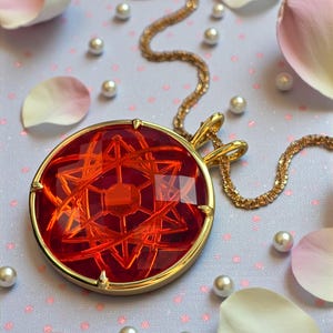 Op de afbeelding: Een goudkleurige hangerketting met een grote, ronde, rode edelsteen. De edelsteen heeft een ingewikkeld, geometrisch ontwerp. De ketting ligt op een lichtpaarse achtergrond met verspreide parels en rozenblaadjes.