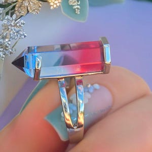 May include: A rectangular ring with a clear, blue, and red crystal. The ring has a silver band and a gold-colored setting around the crystal. The crystal is faceted and has a pointed end.