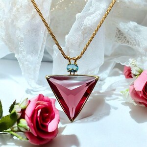 Könnte beinhalten: Eine goldfarbene Halskette mit einem großen, dreieckigen rosa Anhänger und einem kleineren, ovalen blauen Edelstein. Der Anhänger hat einen goldenen Rahmen. Die Halskette wird auf einer weißen Oberfläche mit rosa Rosen präsentiert.