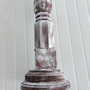 May include: A whitewashed wooden candlestick with a square base and a round top. The candlestick has a distressed finish, giving it a vintage look.