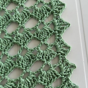May include: Close-up of a handmade, light green crochet doily. The intricate openwork design features a repeating pattern of diamond shapes and a scalloped edge. The yarn has a soft texture, and the color is a muted, natural green.