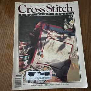 May include: Cross Stitch & Country Crafts magazine cover featuring a white and blue cross stitch sampler with the text "Friendship Memories". The sampler is in a red and white fabric bag with a wooden handle. The bag is sitting on a wooden surface with a glass pitcher of lemonade and a stack of colorful fabric scraps.