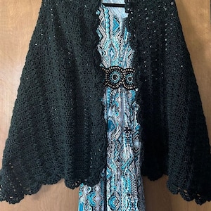 May include: A dark green crochet shawl with a scalloped edge. The shawl is draped over a dress with a turquoise and white geometric pattern.