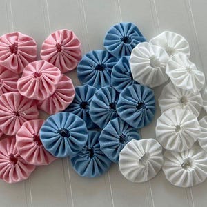 May include: A collection of fabric flowers in three colors: pink, blue, and white. Each flower has a circular shape with pleated petals, creating a textured, decorative element. These could be used for crafting or embellishments.