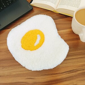 May include: A white fuzzy rug shaped like a fried egg with a yellow yolk in the center. The rug is on a wooden surface.