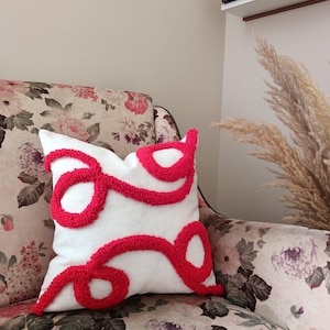 May include: White throw pillow with a red, abstract design. The pillow is made of a soft, textured fabric and has a fluffy, pom-pom like texture.