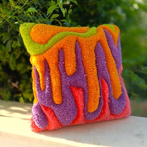 Può includere: Un cuscino quadrato e strutturato con un design astratto vibrante. Il cuscino presenta una combinazione di tonalità arancioni, viola, verdi e rosse, creando un pezzo visivamente sorprendente e artistico. Il design sembra essere un motivo a strati e fluido.