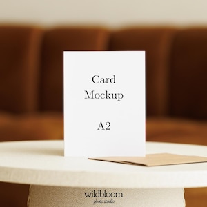 Pode incluir: Uma maquete de cartão branco com o texto "Card Mockup" e "A2" é exibida em uma mesa branca. Um envelope marrom repousa sobre a mesa ao lado do cartão. O fundo apresenta um sofá marrom desfocado. O logotipo do estúdio fotográfico Wildbloom está na parte inferior.