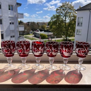 Könnte beinhalten: Sechs rote Weingläser mit klaren Stielen und Böden stehen in einer Reihe. Die Gläser haben ein dekoratives geätztes Design und werfen Schatten. Der Hintergrund zeigt einen blauen Himmel und Gebäude.