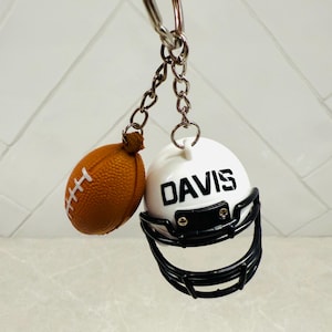 May include: A football-themed keychain featuring a miniature brown football and a white helmet with the name "DAVIS" in black. The helmet has a black facemask. The keychain is attached to a silver chain and ring.