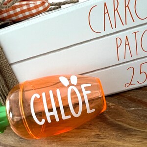 Może przedstawiać: Pomarańczowy plastikowy pojemnik w kształcie marchewki z zielonym wierzchołkiem i napisem "CHLOE" w białych literach. Pojemnik stoi na drewnianej powierzchni przed białym znakiem z napisem "CARROT PATCH. 25 zł"