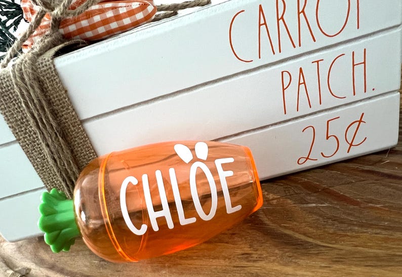 Może przedstawiać: Pomarańczowy plastikowy pojemnik w kształcie marchewki z zielonym wierzchołkiem i napisem "CHLOE" w białych literach. Pojemnik stoi na drewnianej powierzchni przed białym znakiem z napisem "CARROT PATCH. 25 zł"