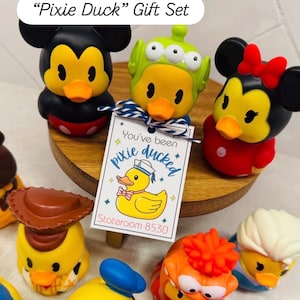 May include: A "Pixie Duck" gift set featuring several character-themed rubber ducks. The ducks are yellow with orange beaks and are styled as Mickey Mouse, Minnie Mouse, an alien, and other characters. A tag reads "You've been pixie ducked."