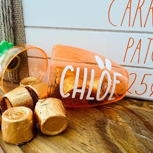Może przedstawiać: Pomarańczowy plastikowy pojemnik w kształcie marchewki z napisem "CHLOE". Pojemnik jest wypełniony małymi, złotymi, owiniętymi w czekoladę cukierkami. Pojemnik stoi na drewnianej powierzchni obok białego znaku z napisem "CARROT PATCH 25&cent;".