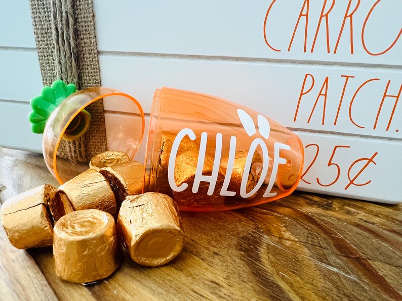 Może przedstawiać: Pomarańczowy plastikowy pojemnik w kształcie marchewki z napisem "CHLOE". Pojemnik jest wypełniony małymi, złotymi, owiniętymi w czekoladę cukierkami. Pojemnik stoi na drewnianej powierzchni obok białego znaku z napisem "CARROT PATCH 25&cent;".
