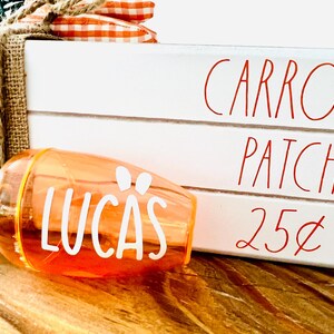 Może przedstawiać: Biały drewniany znak z napisem "Carrot Patch. 25&cent;" w kolorze czerwonym. Przezroczysta pomarańczowa plastikowa butelka w kształcie marchewki z zielonym korkiem i napisem "Lucas" w kolorze białym znajduje się na pierwszym planie.