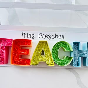 May include: A white box with a clear window displaying the word "TEACH" in colorful, 3D letters. Each letter is a different color: pink, red, yellow-green, and blue. The letters are filled with shredded paper. The name "Mrs. Dreschen" is written above.