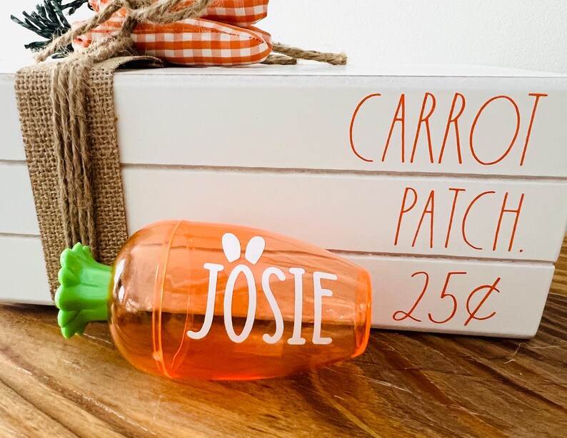 Może przedstawiać: Białe drewniane pudełko z napisem "Carrot Patch 25&cent;". Przed pudełkiem znajduje się plastikowy pojemnik w kształcie marchewki z napisem "Josie".