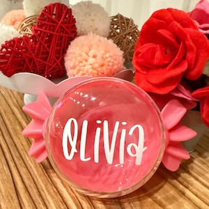 May include: A clear, round ornament with the name "Olivia" in white script. The ornament is filled with pink liquid and has pink bow-shaped accents. Decorative red roses and other ornaments are in the background.