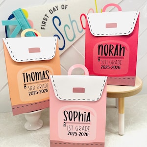 May include: Three colorful, personalized paper backpacks with names and grade levels printed on them. The backpacks are peach, pink, and red, with white flaps and handles. A banner reading "First Day of School" is in the background.