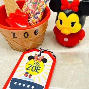 May include: A cruise gift set with a Mickey Mouse-themed toy, a red ice cream bowl with a wooden spoon, and a tag that says "To: Zoe". The set includes a bag of colorful sprinkles.