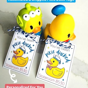 May include: Two yellow and blue toy ducks with attached tags that read "You've been pixie ducked" and "Stateroom 8530". The tags are tied with blue and white twine. The top of the image reads "Assembled & Shipped Pixie Duck Tags".
