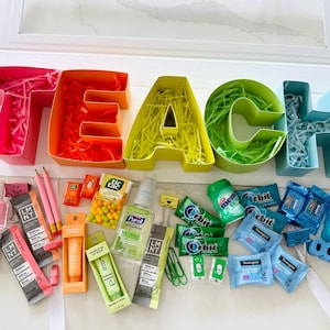May include: A colorful gift box spelling "TEACH" with individual letters in pink, orange, yellow, green, and blue. The letters are filled with shredded paper. Assorted small gifts, including lip balm, gum, and pencils, are arranged below.