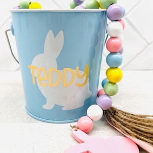 May include: A blue metal bucket with a white bunny silhouette and the word "Teddy" in gold lettering. A pastel colored beaded garland with a pink bunny charm hangs from the bucket.