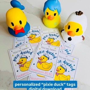May include: A collection of yellow rubber duck toys with blue, white, and black accents, alongside several rectangular tags. The tags feature a cartoon duck wearing a sailor hat and the text "You've been pixie ducked". The bottom text reads "personalized 'pixie duck' tags digital download".