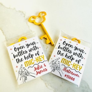 May include: A gold-colored metal bottle opener shaped like a mouse head silhouette, accompanied by two rectangular cards in clear plastic sleeves. The cards feature black text reading Open your bottles with the help of MIC-KEY and illustrations.