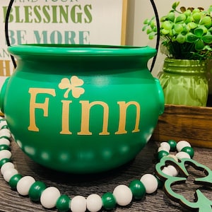 May include: A green pot with the name "Finn" in gold lettering and a shamrock. A black handle arches over the pot. A garland of green and white beads and a wooden shamrock are in the foreground. Decorative plants are in the background.