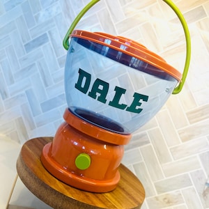May include: A clear plastic gumball machine with the name "DALE" in green lettering. The machine has an orange base and lid, a green handle, and sits on a wooden stand.
