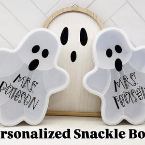 May include: Two white ghost-shaped snack boxes with black eyes and mouths, each inscribed with a name. A wooden ghost cutout is in the background. The text "Personalized Snack Boxes" is at the bottom.