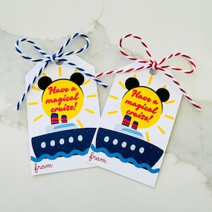 Könnte beinhalten: Zwei weiße Geschenkanhänger mit einem Kreuzfahrtschiff-Design. Die Anhänger zeigen eine gelbe Sonne mit dem Text "Have a magical cruise!" und Mickey Mouse Ohren. Jeder Anhänger hat eine blau-weiße oder rot-weiße Schnur-Schleife.