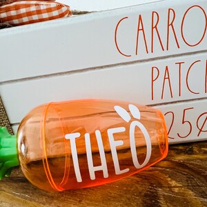 Może przedstawiać: Pomarańczowy plastikowy pojemnik w kształcie marchewki z zielonym wierzchołkiem i napisem "THEO" w białych literach. Pojemnik stoi na drewnianej powierzchni przed białym znakiem z napisem "CARROT PATCH. 25&cent;".