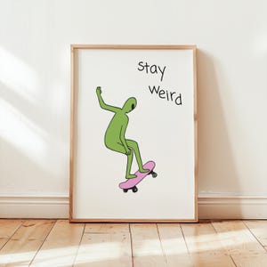 Op de afbeelding: Een groen alienfiguur op een roze skateboard met de tekst "stay weird" in zwarte letters op een witte achtergrond.