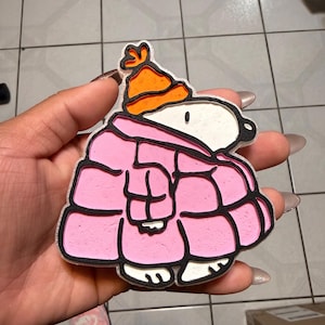 May include: A Snoopy-themed charm in a pink winter coat and orange hat. The charm has a black outline and is held in a hand. The charm is a cartoon character from the Peanuts comic strip.