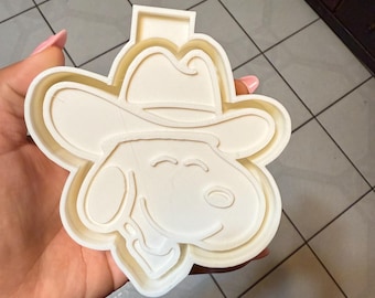 May include: A white plastic cookie cutter in the shape of a cartoon dog wearing a cowboy hat. The cutter has a detailed design with the dog's face and hat clearly visible. The cutter is held in a hand.