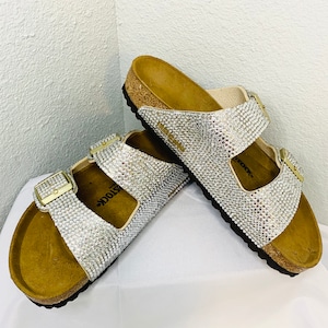 May include: A pair of sparkling silver sandals with a cork footbed and adjustable buckles. The sandals are covered in small, shimmering rhinestones, creating a glamorous look. The buckles are gold-colored.