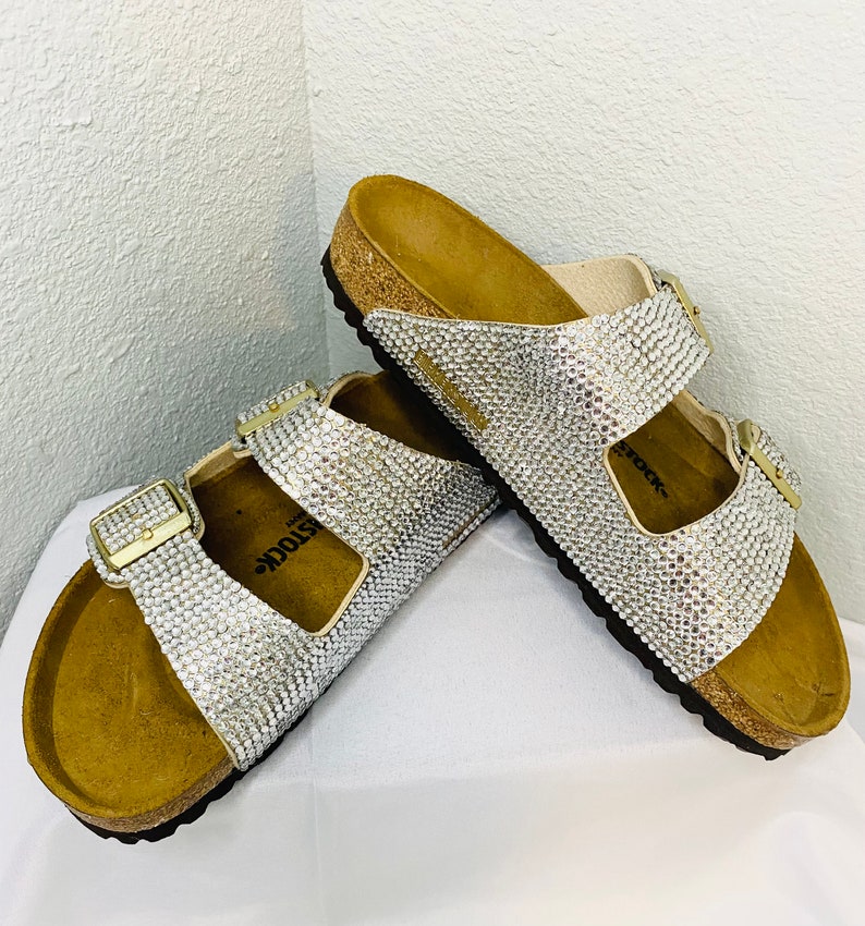 May include: A pair of sparkling silver sandals with a cork footbed and adjustable buckles. The sandals are covered in small, shimmering rhinestones, creating a glamorous look. The buckles are gold-colored.