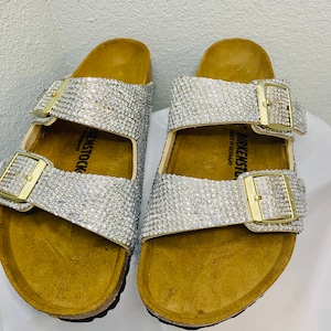 May include: A pair of bedazzled sandals with silver rhinestones covering the straps. The sandals have gold-colored buckles and a cork footbed. The brand name "BIRKENSTOCK" is visible on the side of the sandal.
