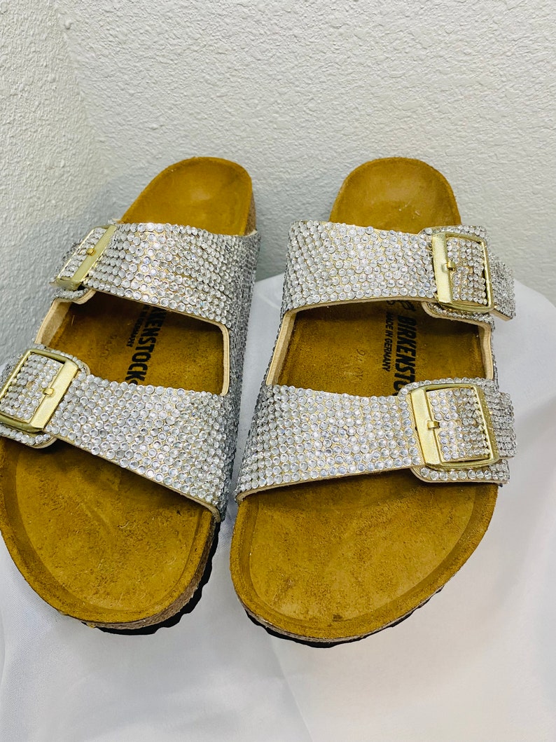 May include: A pair of bedazzled sandals with silver rhinestones covering the straps. The sandals have gold-colored buckles and a cork footbed. The brand name "BIRKENSTOCK" is visible on the side of the sandal.