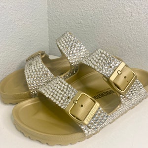 May include: Gold-colored sandals with sparkling rhinestone embellishments. The sandals feature two adjustable straps with gold-tone buckles and the brand name "BIRKENSTOCK" embossed on the buckle. The footbed is contoured for comfort.