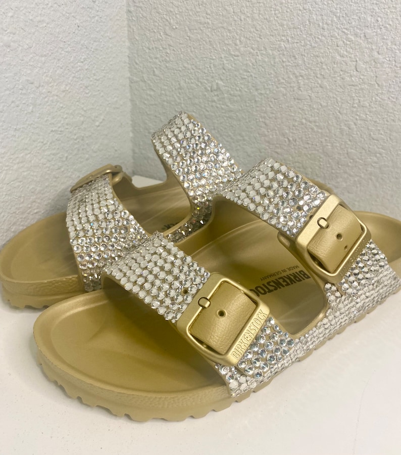 May include: Gold-colored sandals with sparkling rhinestone embellishments. The sandals feature two adjustable straps with gold-tone buckles and the brand name "BIRKENSTOCK" embossed on the buckle. The footbed is contoured for comfort.