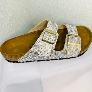 May include: A pair of bedazzled sandals with a cork footbed. The straps and buckles are covered in sparkling rhinestones. The buckles are gold-toned and feature the brand name. The sandals are on a white surface.