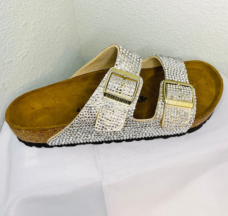 May include: A pair of bedazzled sandals with a cork footbed. The straps and buckles are covered in sparkling rhinestones. The buckles are gold-toned and feature the brand name. The sandals are on a white surface.