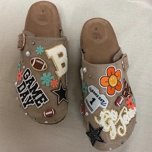 May include: A pair of tan slip-on clogs featuring various patches including a white letter B, a football, a black star, and the text GAME DAY. The other clog displays an orange flower, a jersey patch with the number 1, and the text Go Team.