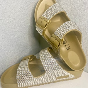 May include: Gold-colored sandals with two straps, each adorned with sparkling rhinestones. The sandals have gold-tone buckles and a contoured footbed. The footwear is designed for comfort and style, suitable for casual wear.